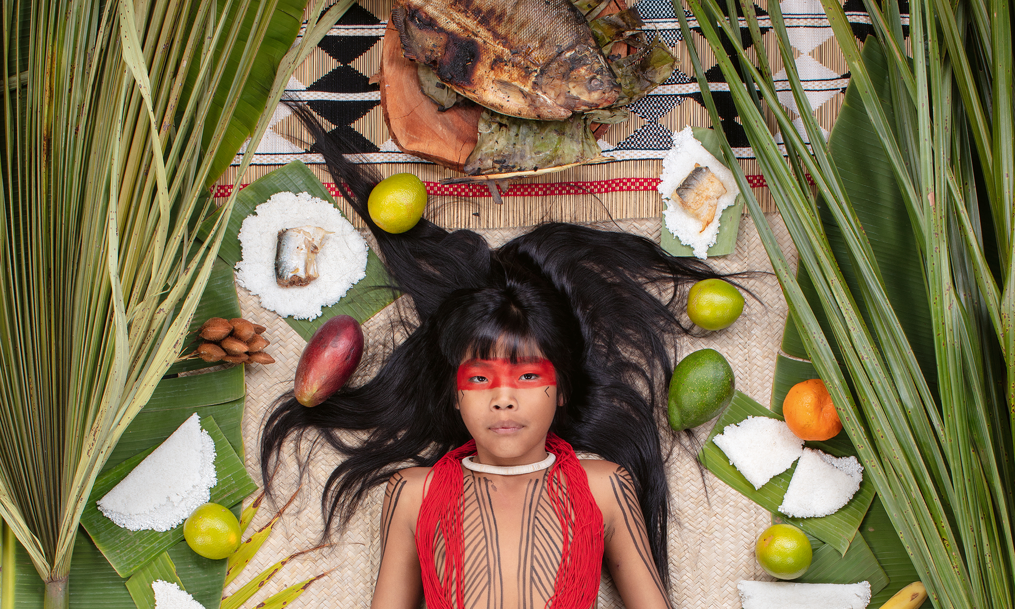 Ein indigenes Kind liegt auf einem Bambustuch, umringt von verschiedenen Speisen, die darstellen, was das Mädchen in einer Woche isst.