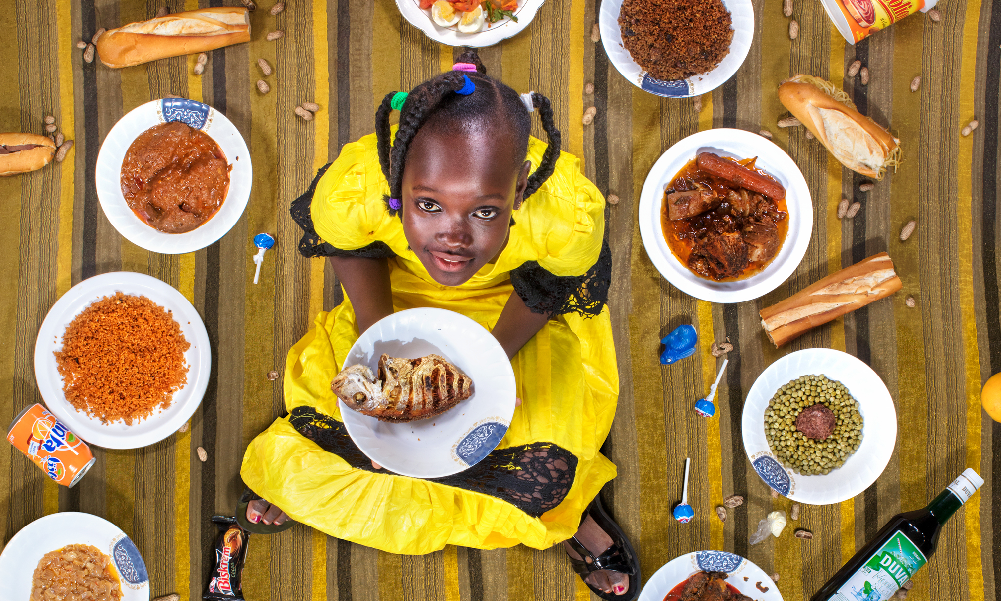Ein senegalesisches Kind sitzt auf einem Teppich, umringt von lokalen Nahrungsmitteln