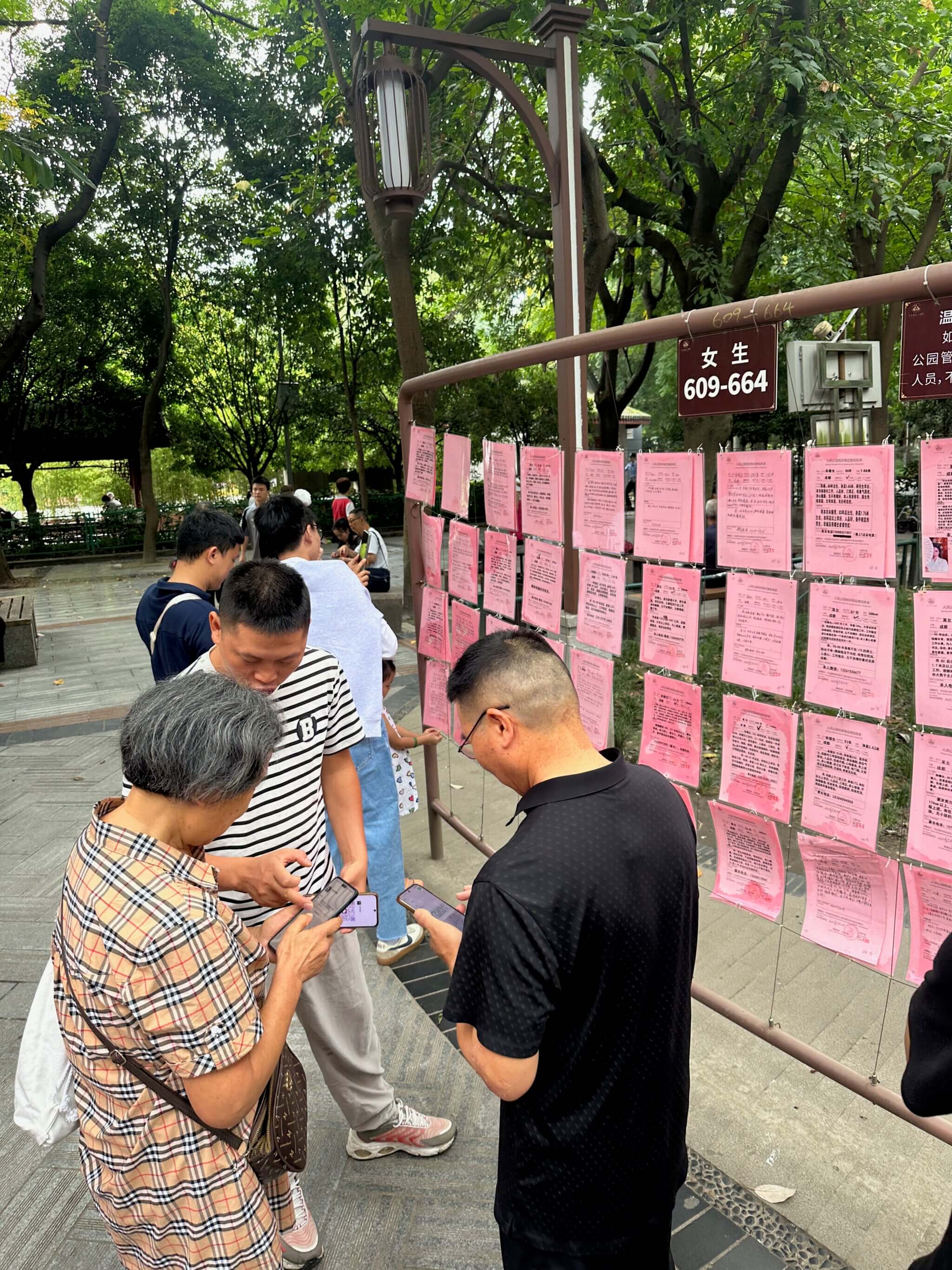 Fünf Menschen, die vor einer Wand stehen, an denen rosa Zettel aufgehängt sind und drei davon tauschen Telefonnummern per Handy aus