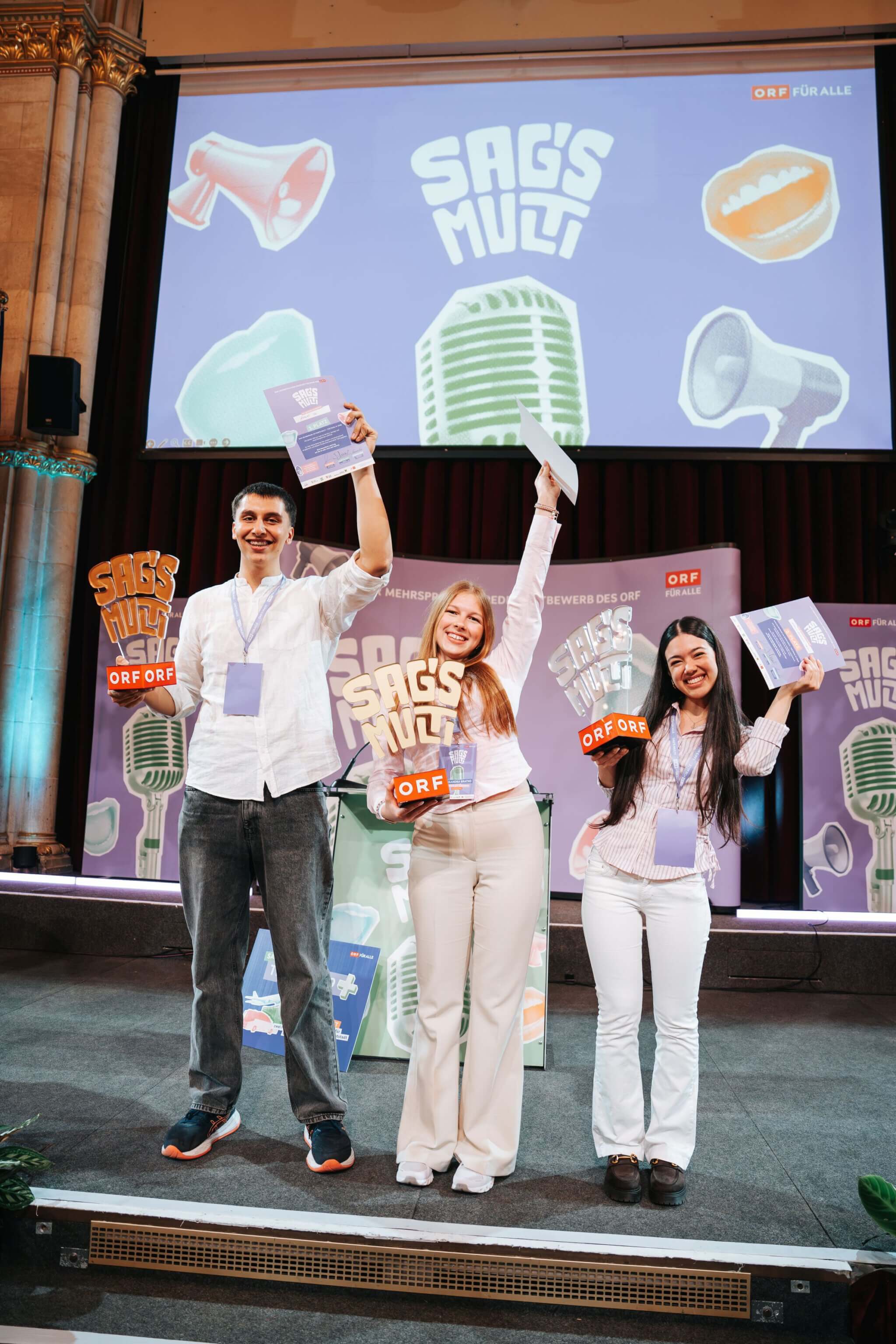 Die 3 Preisträger:innen des Sag´s Multi-Redewettbewerbs 2026 mit ihren Auszeichnungen auf der Bühne im Wiener Rathaus